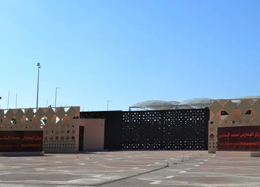 Ein großes Eingangstor mit komplizierten geometrischen Mustern und arabischer Schrift. Im Hintergrund sind moderne Gebäude zu sehen, und der Himmel ist klar und blau.