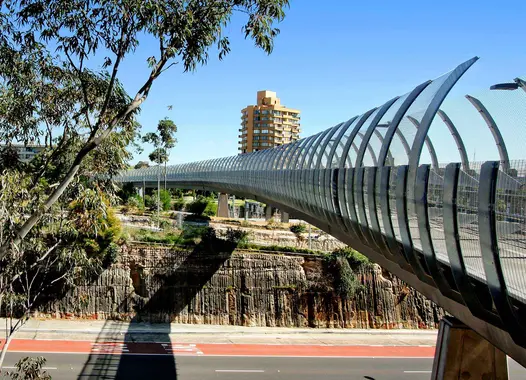 A modern pedestrian bridge with a wave-like roof structure over a street, surrounded by urban scenery under a clear blue sky