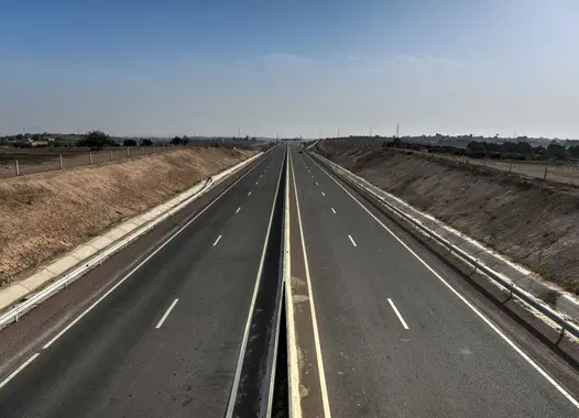 A straight, empty highway with multiple lanes extends into the horizon under a clear sky. The road is flanked by flat, barren land on both sides, with no vehicles or people visible.