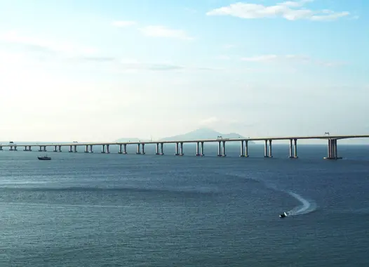 Long bridge with multiple arches spanning across a wide expanse of water under a clear sky, connecting two land masses. Several boats on the water, one creating a wake, with a distant mountain on the horizon.