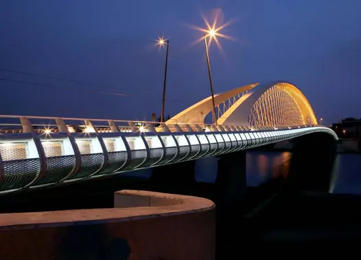 Modern bridge at twilight or night, illuminated by multiple lights. The bridge features a distinctive arching structure with repeating patterns. Starburst light effects are visible, with reflections on the water below.