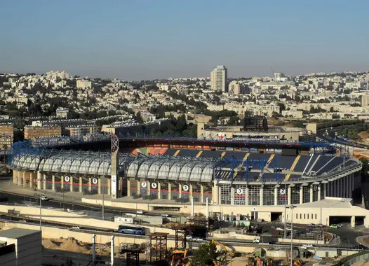 An aerial view of a large stadium with distinctive blue seating and a partial roof structure. Surrounding the stadium is an urban landscape with numerous buildings of varying sizes and designs, roads, and some construction activity in the foreground.