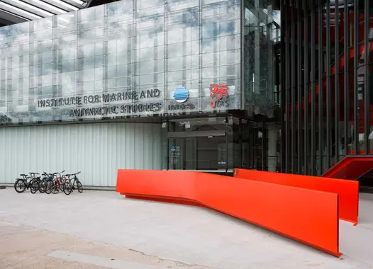 The exterior of a modern building with large glass panels and signage that reads “INSTITUTE FOR MARINE AND ANTARCTIC STUDIES” along with logos next to the text. In front of the building, there is a bright orange, curved bench structure, and to the left side, there are bicycles parked in a bike rack.