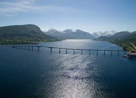 A bridge spans a wide body of water, flanked by green shores and mountains in the background. The shadow of the bridge is clearly cast upon the surface of the water.