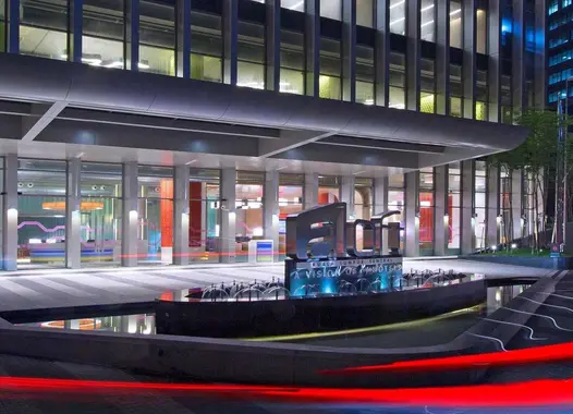 A modern building with a glass facade and a lit-up entrance. Cars with red lights are visible in the foreground, creating a sense of movement.