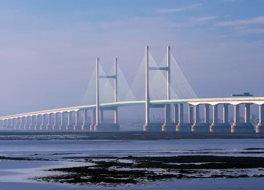 A long, modern suspension bridge with white towers and cables crossing a body of water. The bridge is carrying traffic and the sky is blue.