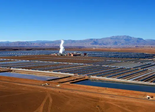 A large-scale solar power plant in a desert landscape. Rows of solar panels are arranged in a grid pattern, with a central tower and steam rising from it. Mountains are visible in the background.