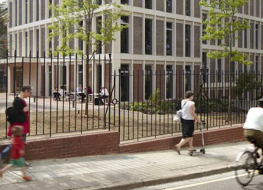 Three-storey building with floor-to-ceiling windows, with people sitting at tables in front of it. Around the house is a courtyard with trees, the house is surrounded by a fence, in front of it people are walking on the pavement and the street. 