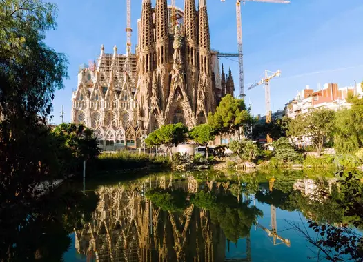 Die Basilika Sagrada Família in Barcelona, Spanien, mit ihren ikonischen Türmen und der komplexen Fassade im Bau, gespiegelt im Wasser darunter.
