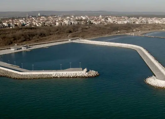 An aerial view of a man-made harbor with a breakwater extending into the sea. The harbor is surrounded by a coastal town and a large body of water.