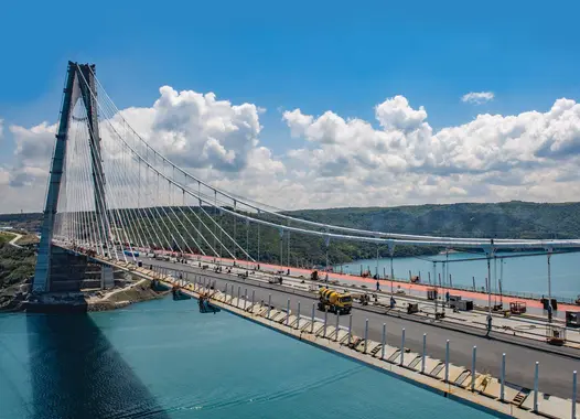 A large suspension bridge under construction, spanning a body of water. The bridge deck is partially completed, with workers and construction vehicles visible.