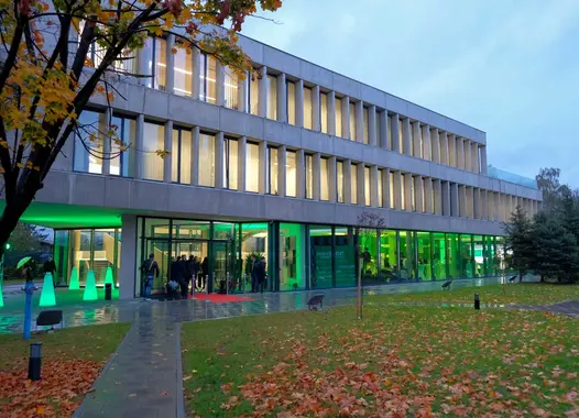 Ein modernes Bürogebäude mit einer Glasfassade. Vor dem Gebäude befinden sich Bäume und ein Gehweg.