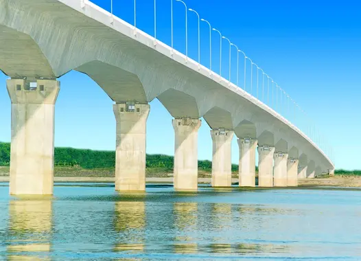 A long bridge with multiple arches spans over a river. The bridge has a series of identical, evenly spaced supports and a flat roadway on top with railings on either side.