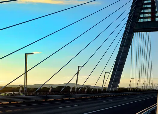 A modern cable-stayed bridge with a white tower and numerous cables stretching from it to the road deck. The sun is setting in the background, casting a warm glow on the bridge.