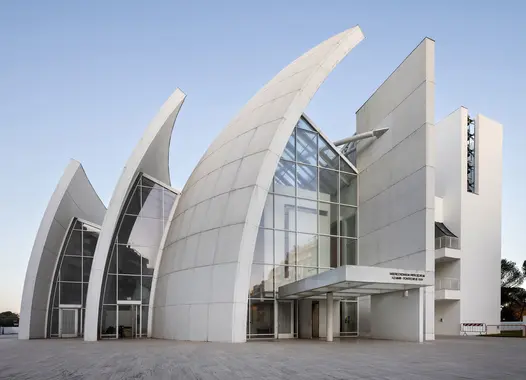 Moderne weiße Kirche mit segelartig geschwungenen Paneelen, die aus dem Boden ragen, einer Glasfassade in der Mitte und einem minimalistischen Design; auf einer gepflasterten Freifläche unter einem klaren blauen Himmel gelegen.