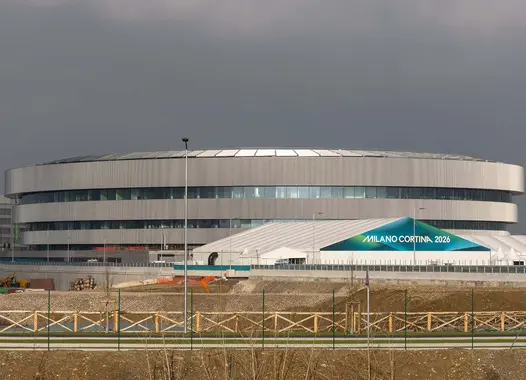 Rundes Gebäude mit metallischer Fassade im Hintergrund, umgeben von weiteren Bauten, darunter ein grünes Zelt mit der Aufschrift „MILANO-CORTINA 2026”.