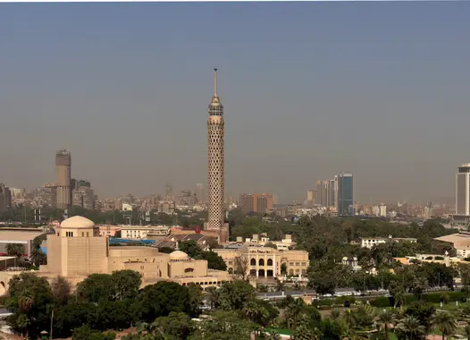 A panoramic view of a city skyline with a prominent tower on the left, surrounded by low-rise buildings and lush greenery under a clear sky.